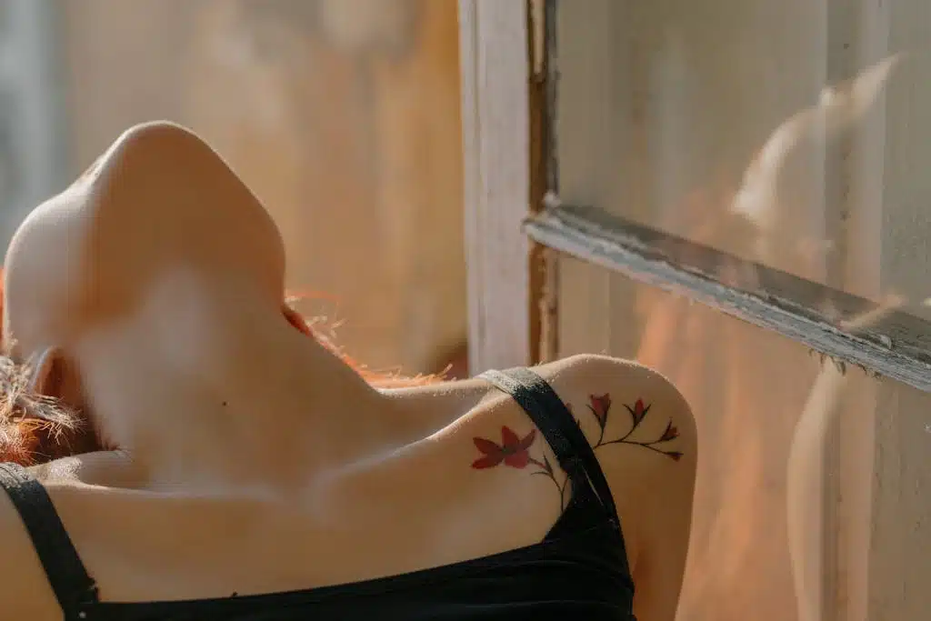 A serene woman leans back in the sunlight, revealing a floral tattoo against her skin.