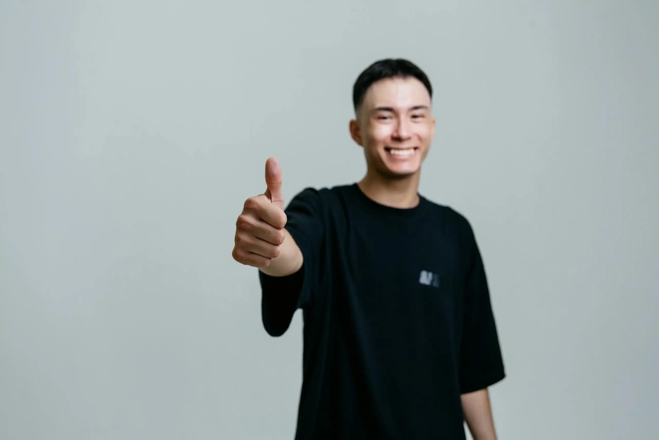 A cheerful young man in a black shirt smiles and gives a thumbs up in a studio setting.