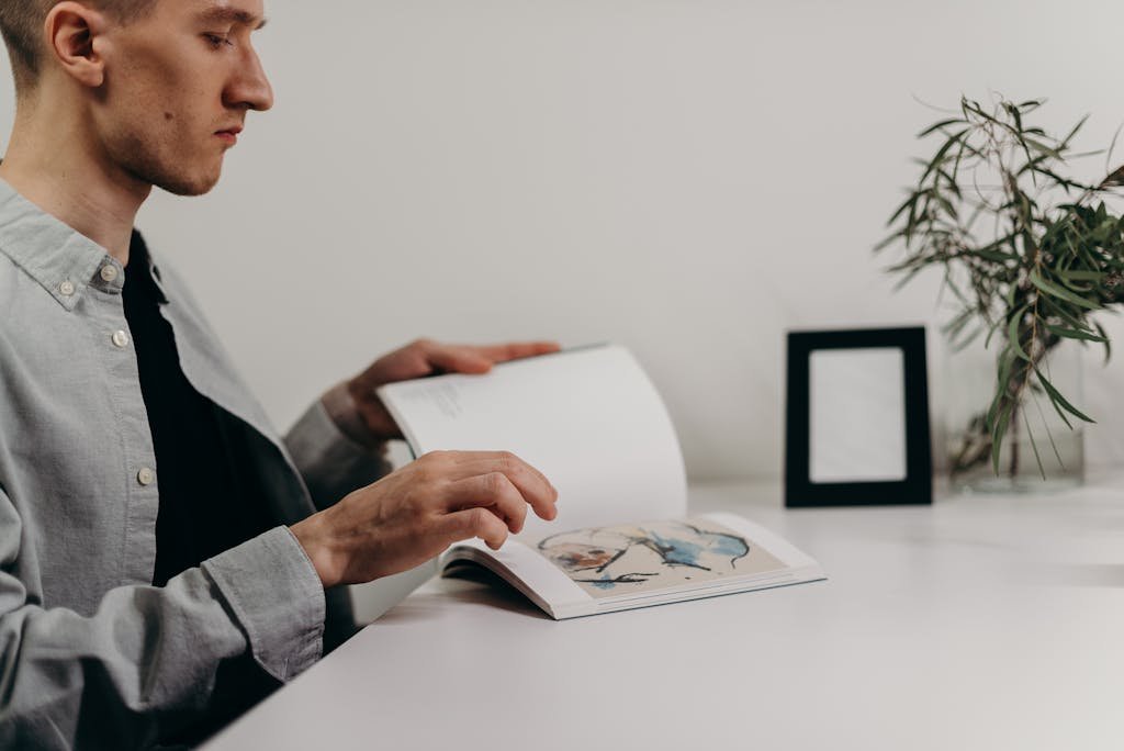 A man reading a book at a minimalistic home office with a plant and black photo frame.
