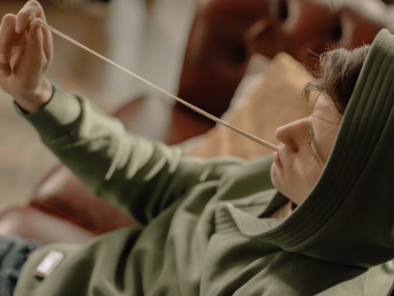 A young man in a green hoodie stretches gum while relaxing on a sofa indoors.