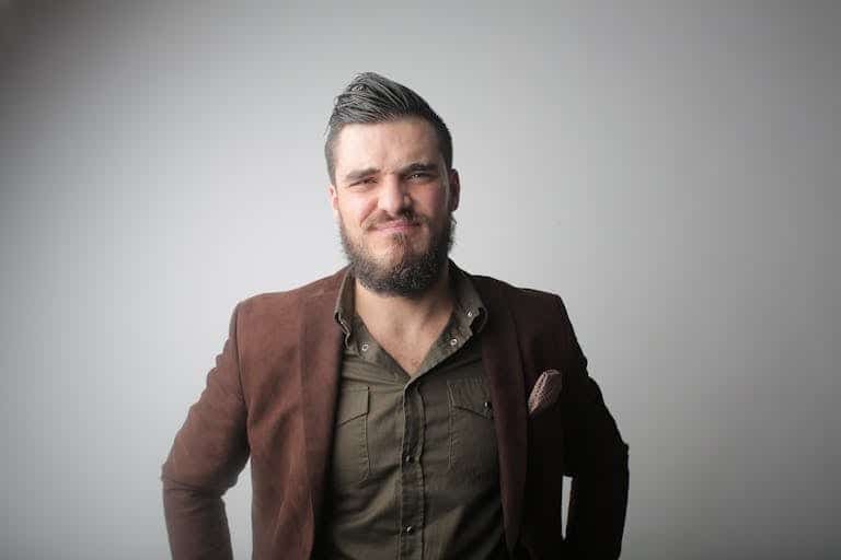 A young man with a full beard poses confidently in stylish outerwear against a gray background.