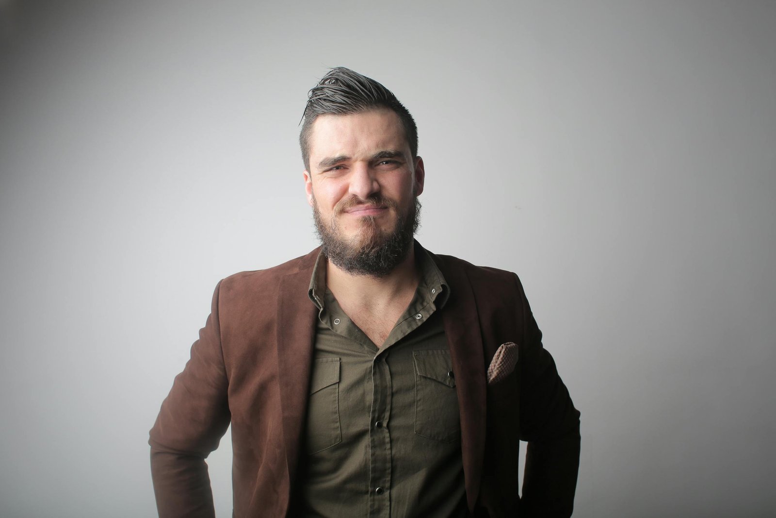 A young man with a full beard poses confidently in stylish outerwear against a gray background.