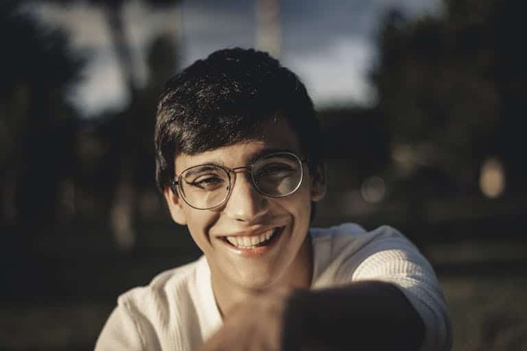 A young man with glasses smiling warmly in a sunlit outdoor setting.