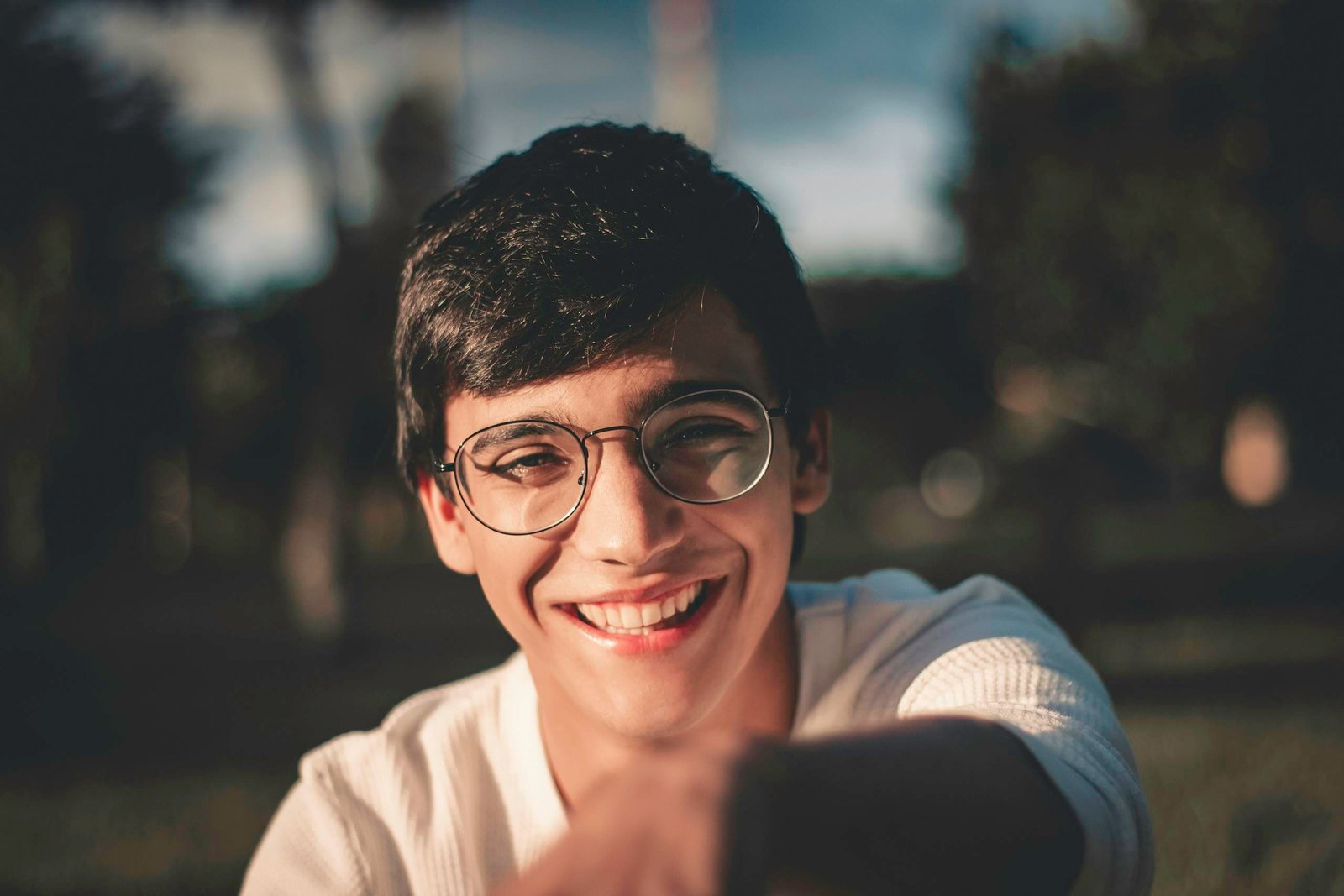 A young man with glasses smiling warmly in a sunlit outdoor setting.