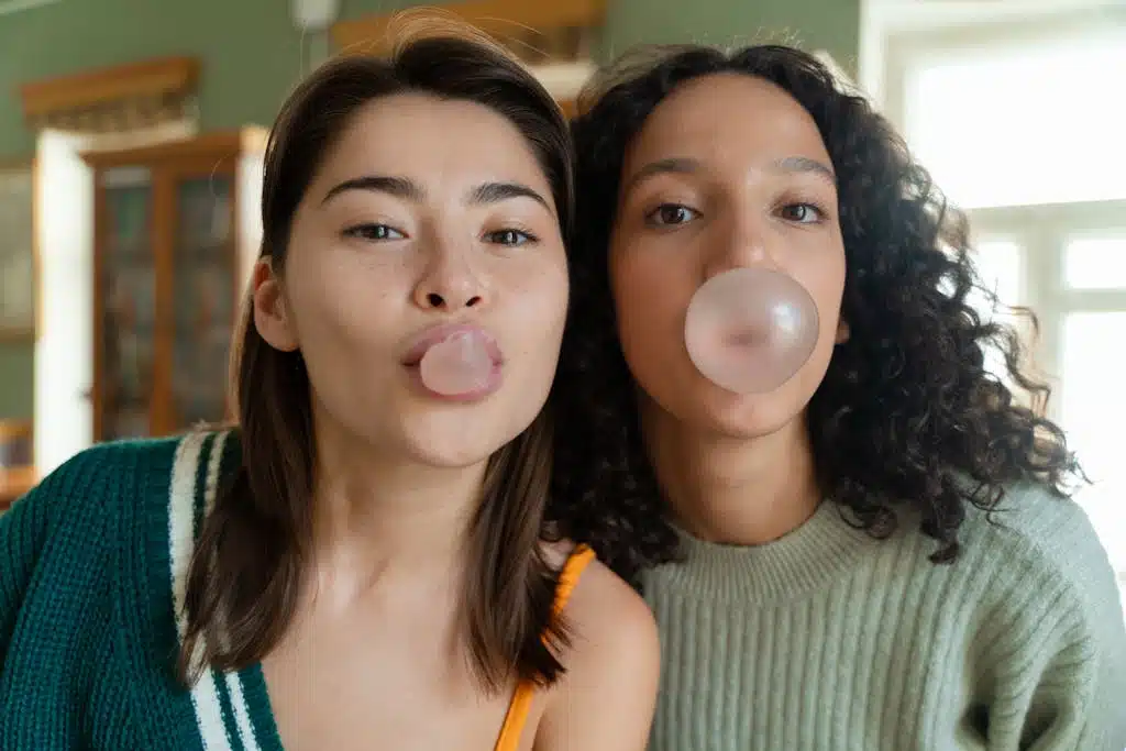 Close-up of two young women blowing bubble gum together indoors, sharing a joyful moment.