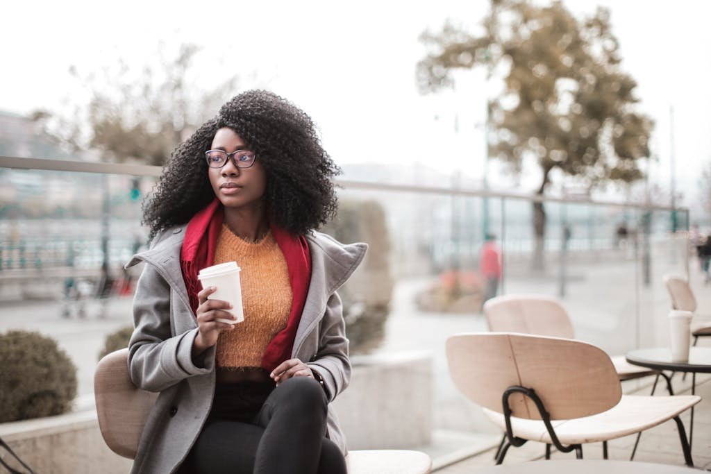 A fashionable woman with glasses enjoys coffee outside on a cool day.