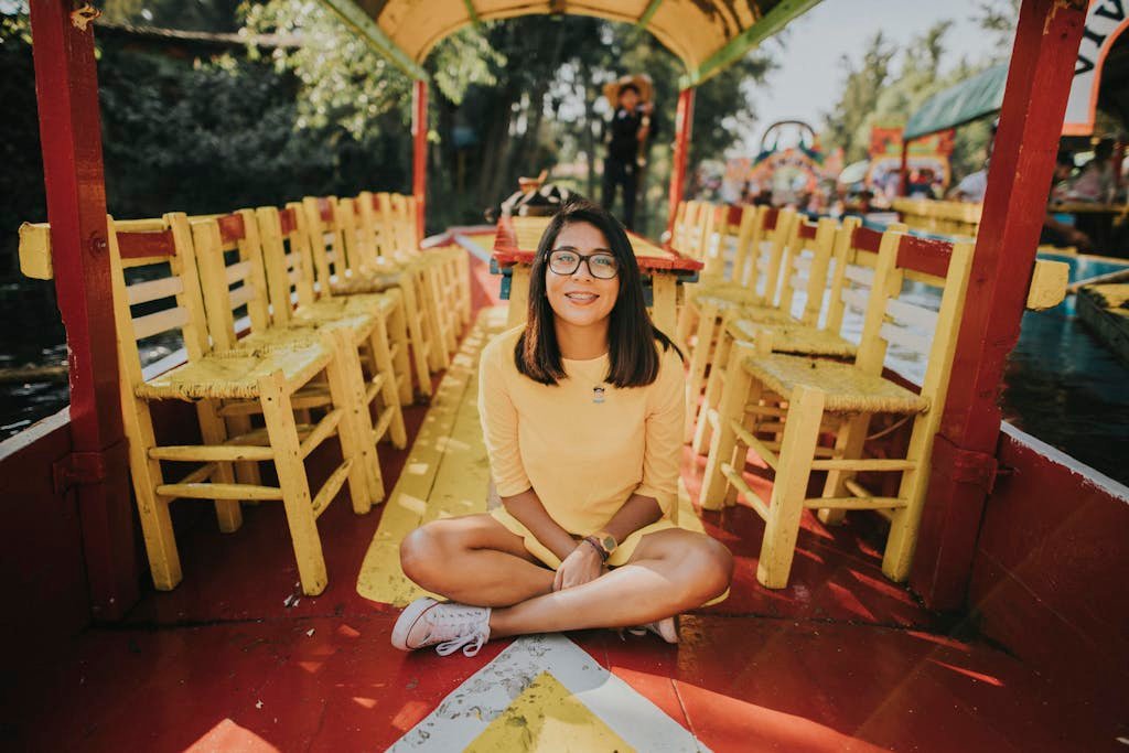 A cheerful woman in a yellow dress sits on a colorful wooden boat surrounded by vibrant chairs in daylight.