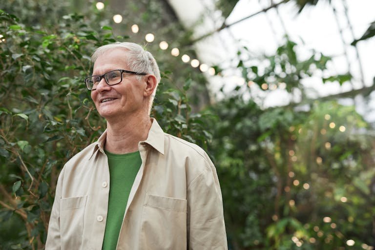 Elderly man smiling warmly inside a greenhouse surrounded by lush greenery.