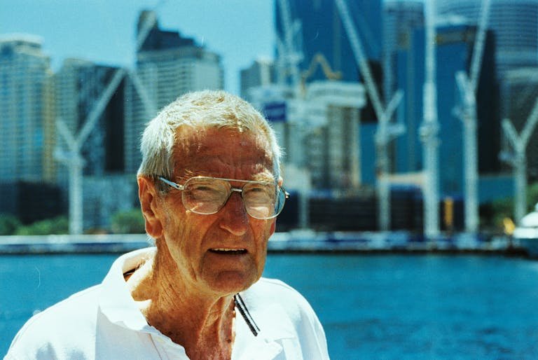 Elderly man wearing glasses at Sydney Harbour with skyscrapers in the background.