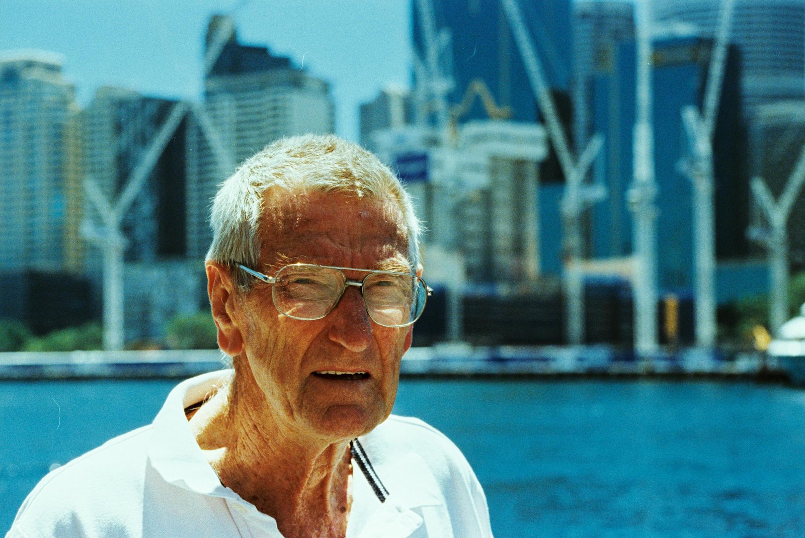 Elderly man wearing glasses at Sydney Harbour with skyscrapers in the background.