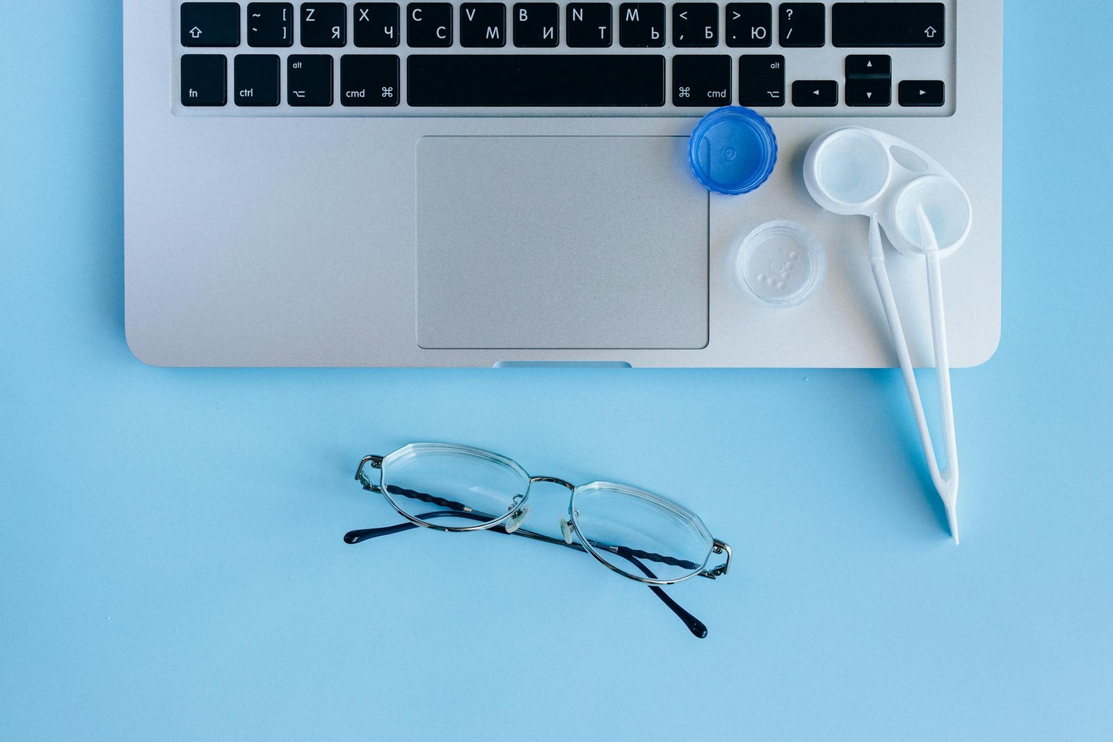 Top view of a laptop keyboard with eyeglasses and contact lens case on a blue background.